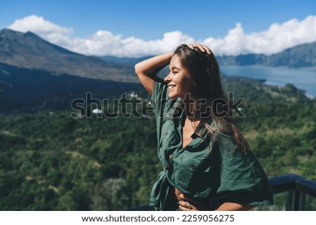 Side view of young cheerful female tourist with long dark hair in casual clothes looking away while resting on terrace of scenic viewpoint against mountain ridge and blue sky Royalty-Free Stock Photo #2259056275