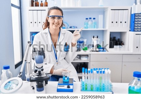 Young hispanic woman working at scientist laboratory with a big smile on face, pointing with hand and finger to the side looking at the camera. 