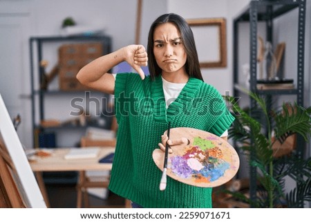 Young south asian woman holding painter palette with angry face, negative sign showing dislike with thumbs down, rejection concept 