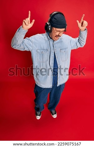 Top view. Portrait of man in stylish modern clothes listening to music in headphones over red background. Concept of modern fashion, lifestyle, music culture, emotions, facial expression. Ad