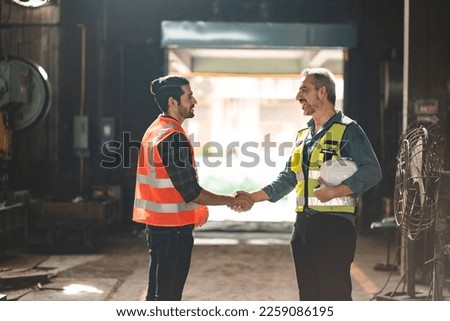 Portrait of senior and young male engineers and workers wearing safety vests and jacket while holding hardhat with arm around shoulder standing in front of machine in warehouse looking at camera Royalty-Free Stock Photo #2259086195