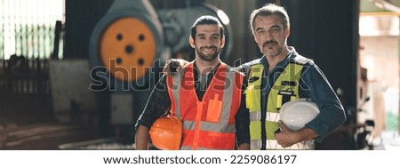 Portrait of senior and young male engineers and workers wearing safety vests and jacket while holding hardhat with arm around shoulder standing in front of machine in warehouse looking at camera Royalty-Free Stock Photo #2259086197