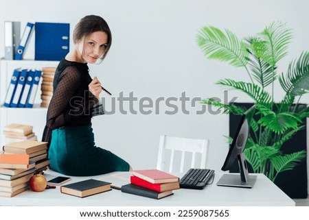 woman in the office sits on a desk near a computer and books