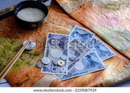 Coins and paper banknotes of Japanese yen, and a bowl of rice and chopsticks on the background of national fabric with embroidery