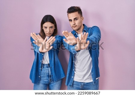 Young hispanic couple standing over pink background rejection expression crossing arms and palms doing negative sign, angry face 