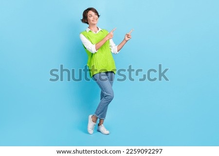 Full size photo of adorable girl with brown hairdo wear green vest staring indicating look empty space isolated on blue color background
