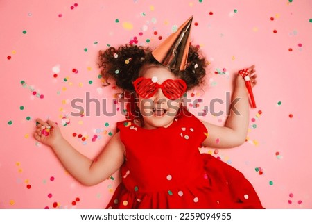 happy little girl in a birthday cap lies among the multicolored confetti on a pink background, a view from above.