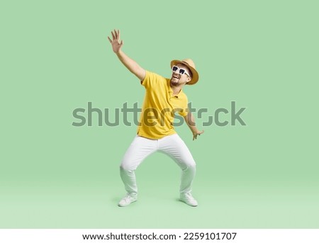 Funny man in casual summer clothes dancing on light green studio background. Happy young traveler wearing yellow tee shirt, white pants, sun hat and sunglasses dancing and having fun on holiday