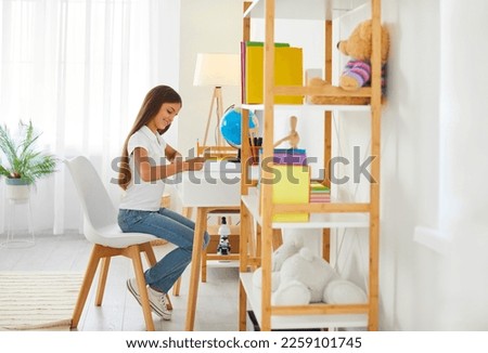 Happy child studying at home. Little kid sitting at a desk in a modern interior. Happy school girl sitting at a table in her beautiful, cozy room, doing homework, writing in her notebook and smiling Royalty-Free Stock Photo #2259101745
