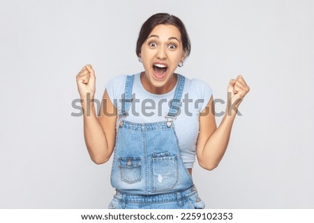 I am champion. Portrait of victorious delighted woman wearing denim overalls clenching fists, shouting for joy, screaming celebrating win success. Indoor studio shot isolated on gray background. Royalty-Free Stock Photo #2259102353