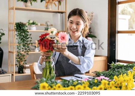 Female florist in apron smiling and arranging gerbera flowers in glass vase with enjoying while creating and designing floral bouquet in her flower shop. Royalty-Free Stock Photo #2259103095