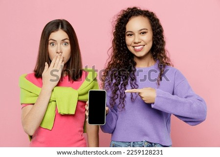 Young two friends women wear green purple shirts together hold in hand use point index finger on mobile cell phone with blank screen workspace area isolated on pastel plain light pink color background