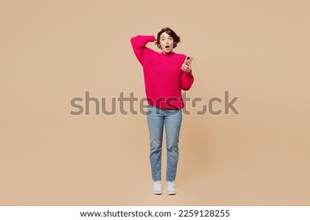 Full body young sad confused puzzled woman wearing pink sweater holdhead using mobile cell phone look camera isolated on plain pastel light beige background studio portrait. People lifestyle concept Royalty-Free Stock Photo #2259128255