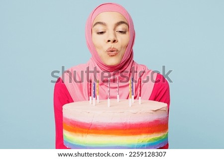 Young happy fun arabian muslim woman wearing pink abaya hijab holding cake blow out candles making wish isolated on plain pastel light blue cyan background studio. People uae islam religious concept