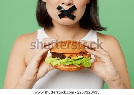 Cropped young wistful woman with mouth sealed with tape wear white clothes hold look at burger isolated on plain pastel green background. Proper nutrition healthy fast food unhealthy choice concept