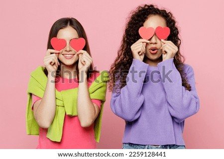 Young two friends fun cheerful women 20s wears green purple shirts together hold in hand red paper hearts cover eyes with stickers isolated on pastel plain light pink color background studio portrait
