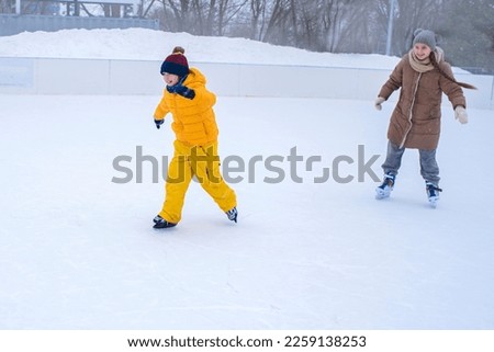 A little boy in yellow clothes is skating with his older sister a teenager in winter at a street skating rink