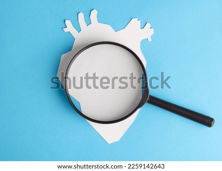 Anatomical heart cut out of white paper with a magnifying glass on a blue background. Healthy heart concept