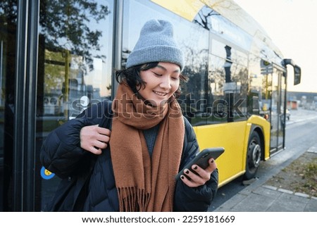 Image of girl student waiting for public transport, checks schedule on smartphone app, stands near city bus. Urban lifestyle concept