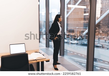Full body side view of businesswoman in formal clothes standing near window while looking through glass wall in modern office Royalty-Free Stock Photo #2259182761