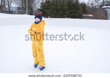 happy boy in bright yellow winter clothes in hockey skates stands on the ice rink alone outside the winter