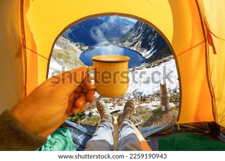 Cup of hot drink in the hand and wonderful mountain lake through the open entrance of the tent. The beauty of a romantic hike and camping accompanied by a dog.
