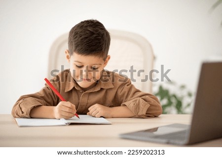 Smiling cute small european kid sitting at table studying, drawing with laptop, enjoy lesson at school in room interior. Art, learning at home with device, childhood and elementary education remotely