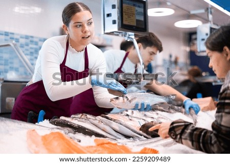Smiling salesgirl in work clothes holding in hands fresh seabass and offering it to purchaser in fish shop Royalty-Free Stock Photo #2259209489