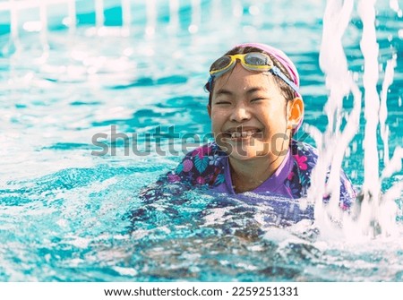 Portrait of a happy and healthy Asian child girl with goggles swimming in a swimming pool, big smiling, eyes looking at the camera, space for text and design.