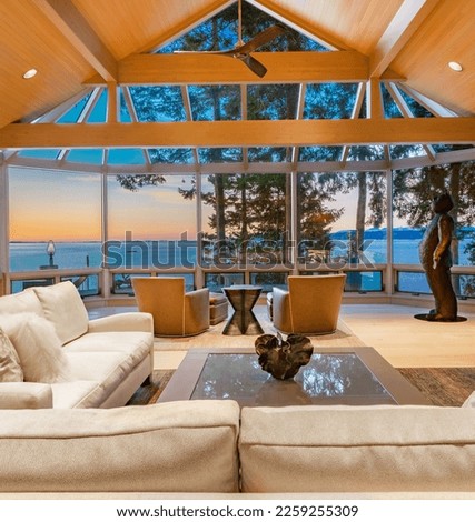 stately and elegant living room of a waterfront home interior space with wall of windows and sky lights with bright colourful skies at sunset and dusk blue hour rich leather chairs fireplace Royalty-Free Stock Photo #2259255309