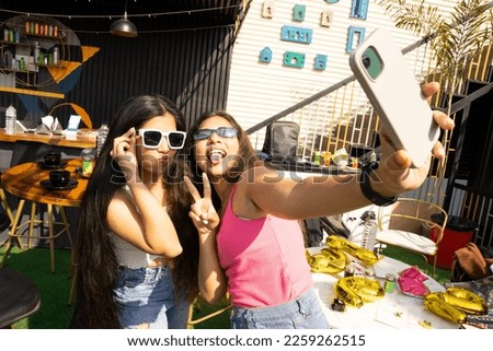 Two joyful cheerful young indian girls taking a selfie together at cafe and showing peace gesture and making funny faces outdoors