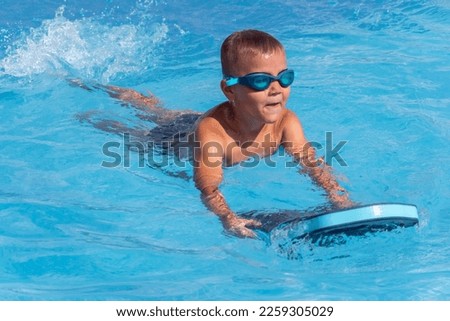 Happy boy child in swimming goggles with float foam board tool swim in pool in sunny day. Safe pool training, summer holiday, family vacation, travel, water sport concept Royalty-Free Stock Photo #2259305029