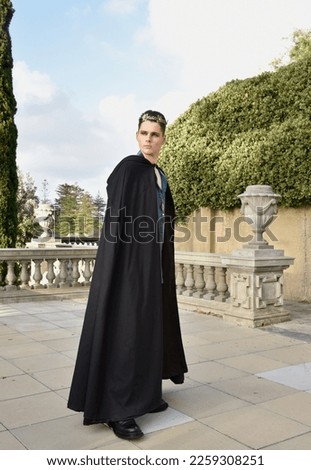 portrait of handsome brunette man wearing fantasy medieval prince costume with golden crown and romantic silk shirt and dark flowing black cloak. historical castle location background. Royalty-Free Stock Photo #2259308251