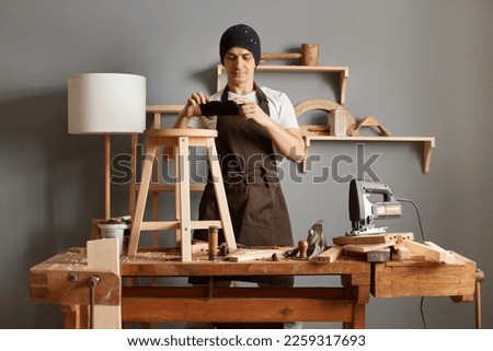 Portrait of a young male carpenter in a black hat and brown apron posing in his carpentry workshop, taking a photo on the phone of his product, making wooden chairs.