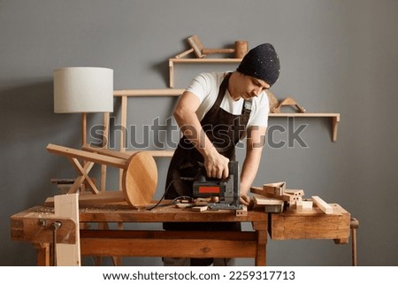 Indoor shot of man carpenter wearing white t-shirt, brown apron and black cap, working on woodworking machines in carpentry shop, sawing a plywood sheet with jig saw machine. Royalty-Free Stock Photo #2259317713