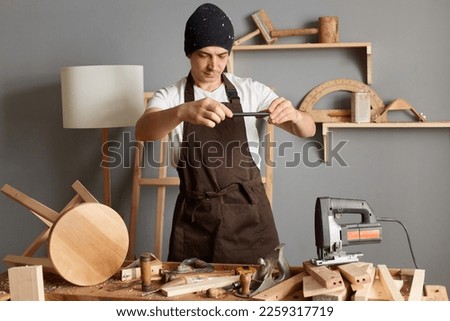 Indoor shot of Caucasian handsome carpenter in a black hat and brown apron working workshop, taking a photo on mobile phone of his product, posing in joinery.