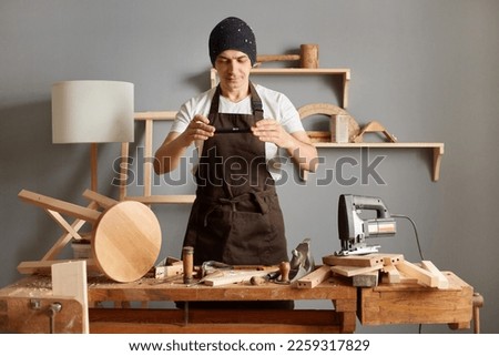 Indoor shot of handsome young adult man carpenter wearing brown apron and black cap working in his carpentry workshop, making creative wood products, photographing his work.