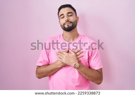 Hispanic young man standing over pink background smiling with hands on chest with closed eyes and grateful gesture on face. health concept. 