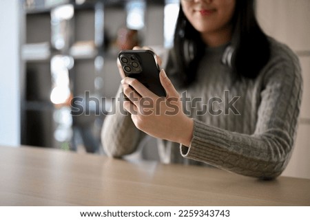 Pretty Asian woman in cozy sweater sits at the desk using her smartphone, chatting with her friends, scrolling on online shopping app. cropped image
