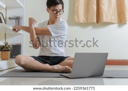 Heathy Asian man stretching shoulder, using laptop for learning yoga stretching for relax on online yoga class at home. Man stretching before workout at home. Home online workout yoga exercise concept Royalty-Free Stock Photo #2259361771