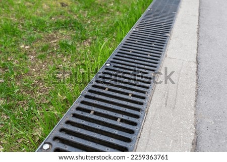 A rusted metal sewer grate along a paved walkway. The edge of the pavement. Close-up, diagonal