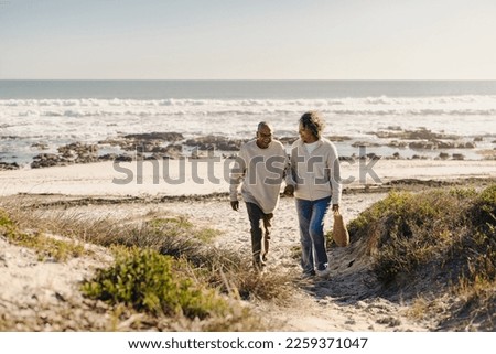 Happy senior couple smiling while walking away from the beach after a romantic picnic. Cheerful elderly couple enjoying a seaside holiday after retirement. Royalty-Free Stock Photo #2259371047