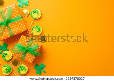 Saint Patrick's Day concept. Top view photo of gift boxes with green ribbon bows meringue lollipops sprinkles gold coins and trefoils on isolated orange background with empty space