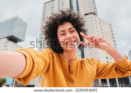 Young hispanic woman with curly hair, yellow shirt and cheerful attitude, smiling and having fun taking a selfie photo doing the peace sign with the fingers at city street outdoors. Lifestyle concept