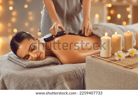 Woman enjoying exotic hot stones spa massage. Relaxed young woman lying on a spa bed while the masseuse is putting hot stones on her back. Spa treatment concept Royalty-Free Stock Photo #2259407733