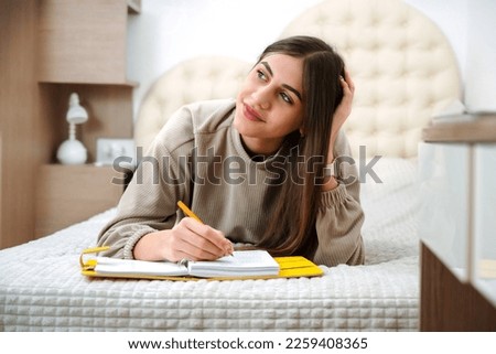 Teen girl lies on the bed and writes in a notebook, keeps a diary book, diary planner. Royalty-Free Stock Photo #2259408365