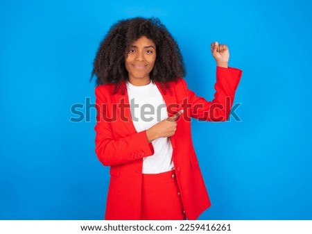 Smiling young businesswoman with afro hairstyle wearing red over blue wall raises hand to show muscles, feels confident in victory, strong and independent.
