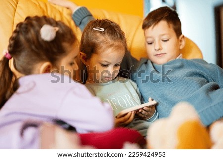 Сhildren sit on a leather sofa in the living room and watch cartoons on phone.