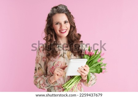 happy stylish 40 years old woman in floral dress with tulips bouquet and tablet PC isolated on pink.