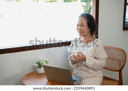 Elderly women sit and relax, stressed, watching series on laptops, reading and taking notes in notebooks and sipping tea happily inside the house with the brightest light and fresh air. 